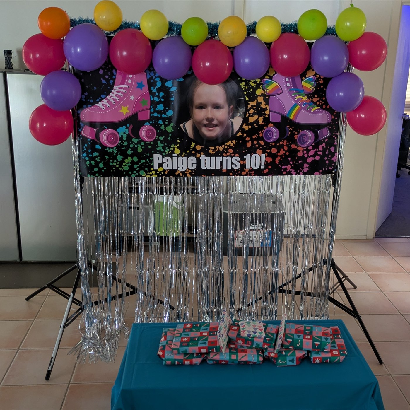 Finished Paige turns 10 personalised photo birthday banner displayed on a backdrop stand with silver fringe and colourful balloons