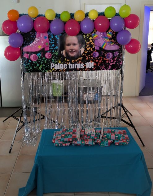 Finished Paige turns 10 personalised photo birthday banner displayed on a backdrop stand with silver fringe and colourful balloons
