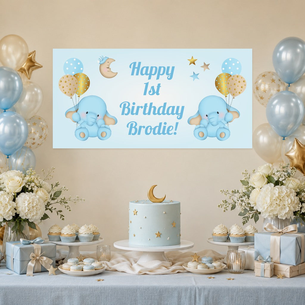 Blue and gold elephant first birthday decorations with Brodie's personalised banner, a pale blue moon and stars cake, cupcakes and balloons.